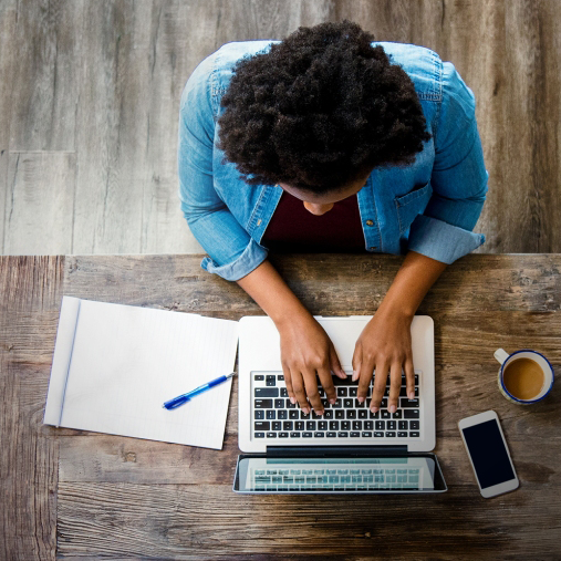 A photo from above of a person typing on a laptop with a phone, notepad, pen, and mug of coffee beside them