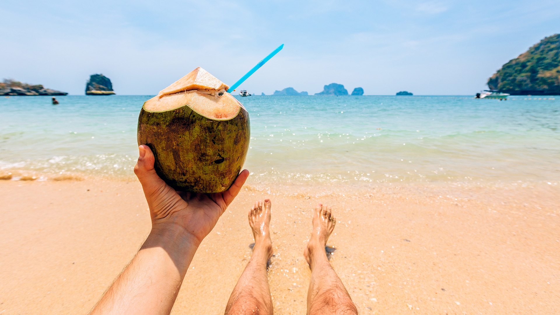 Man drinking fresh coconut milk while relaxing by the ocean at the beach, personal perspective view - stock photo